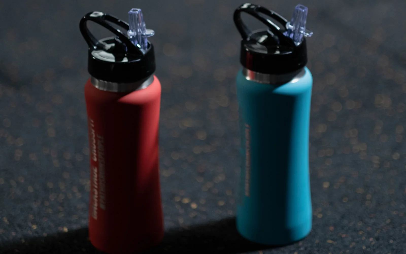 A red and a blue sports water bottle on a dark gym floor.